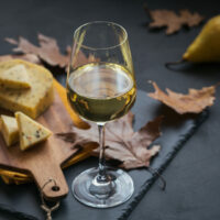 A glass of white wine was served with cheese in a cutting board on dark background. Autumn picnic with cheese, wine and dry leaves in rustic style.