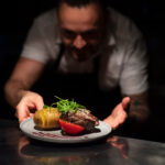 Chef handing plate with meal through order station in the commercial kitchen on black background.